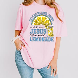 A woman wearing a pink t-shirt with a graphic design of a lemonade glass and text that says "Life gives me some lemons but my JESUS He be makin' lemonade".