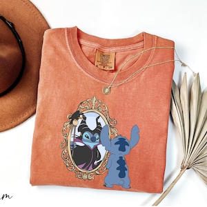 a red t-shirt with a graphic of two cartoon characters, a blue one and a black one, standing in front of a mirror. The t-shirt is placed on a white surface, along with a brown hat, a necklace, and a fan.