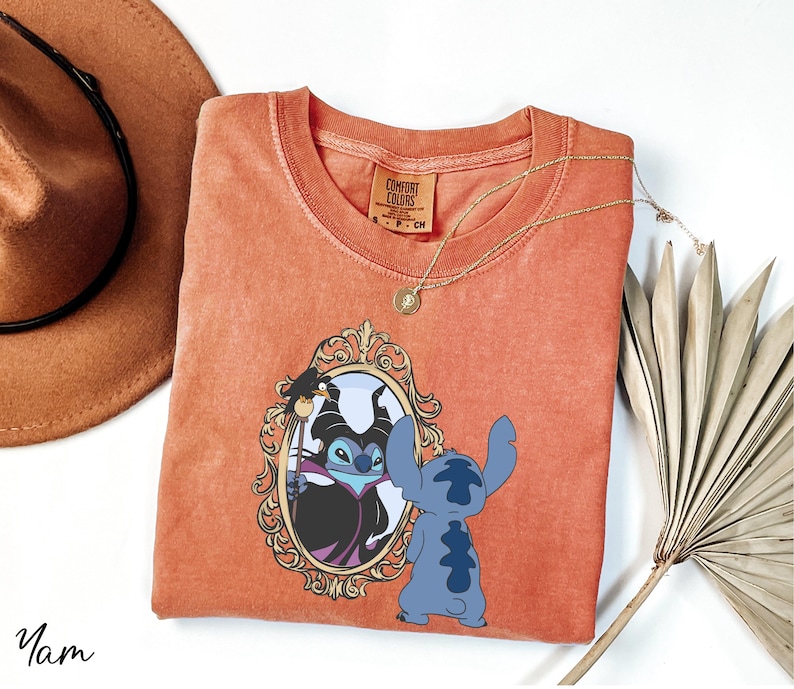 a red t-shirt with a graphic of two cartoon characters, a blue one and a black one, standing in front of a mirror. The t-shirt is placed on a white surface, along with a brown hat, a necklace, and a fan.