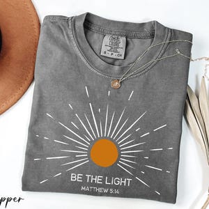 K&ouml;nnte beinhalten: Graues T-Shirt mit einem wei&szlig;en Sonnenstrahlen-Design und dem Text "Sei das Licht" mit dem Bibelvers "Matth&auml;us 5:14".