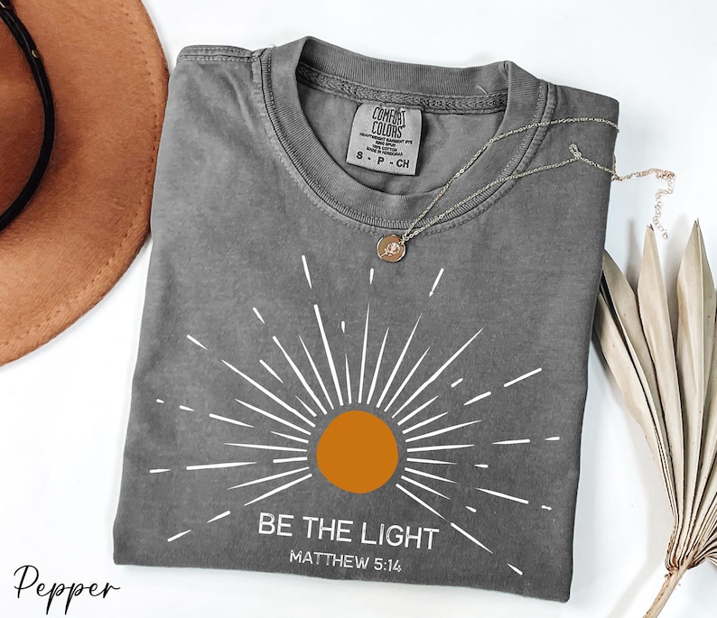 K&ouml;nnte beinhalten: Graues T-Shirt mit einem wei&szlig;en Sonnenstrahlen-Design und dem Text "Sei das Licht" mit dem Bibelvers "Matth&auml;us 5:14".