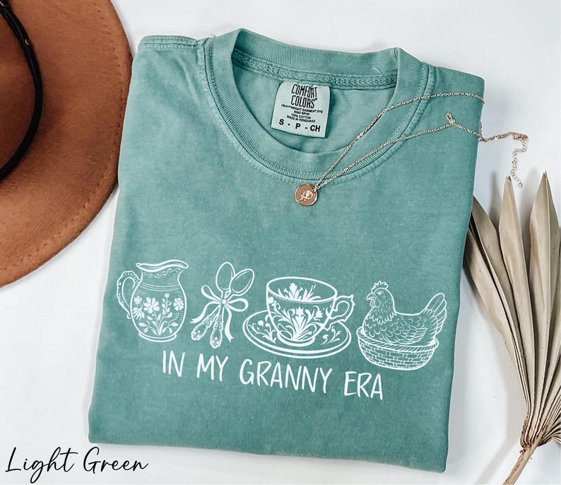 A light green t-shirt with a graphic design of kitchen utensils and a chicken, accompanied by a necklace and a brown hat.
