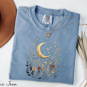 A blue denim t-shirt with a moon and starry night sky design, along with a necklace and a brown hat, are arranged on a white surface.