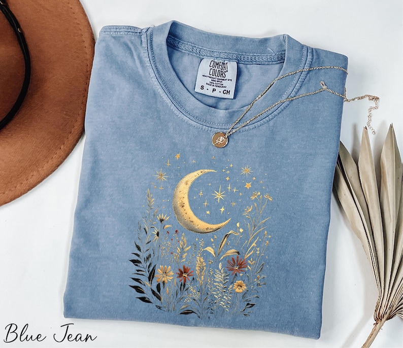 A blue denim t-shirt with a moon and starry night sky design, along with a necklace and a brown hat, are arranged on a white surface.