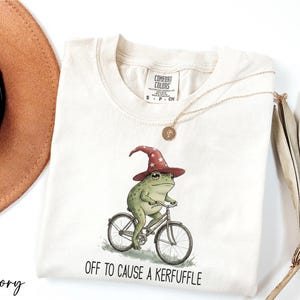 A white t-shirt with a whimsical illustration of a frog riding a bicycle, wearing a wizard's hat, and the text "Off to cause a Keruffuffle" below the image.