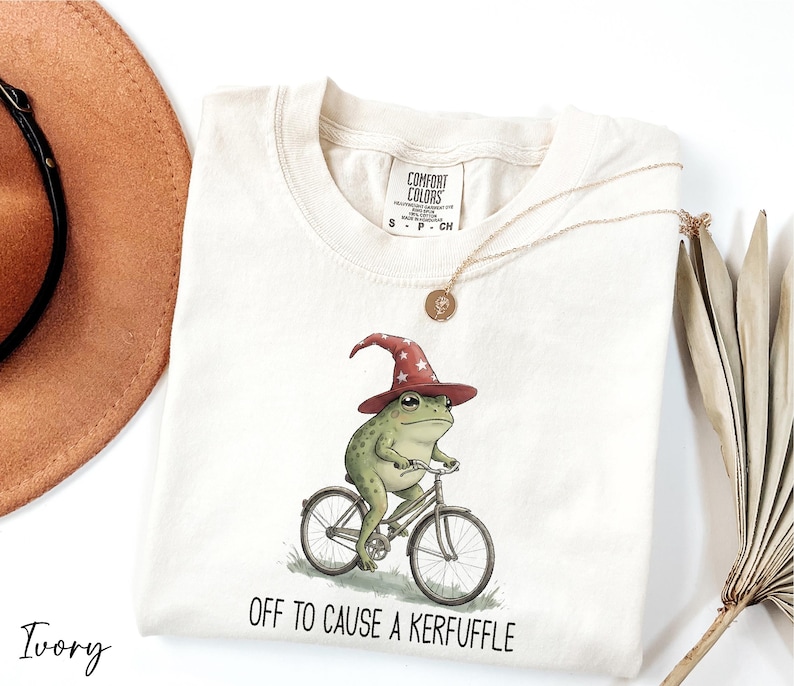 A white t-shirt with a whimsical illustration of a frog riding a bicycle, wearing a wizard's hat, and the text "Off to cause a Keruffuffle" below the image.