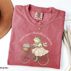 a red t-shirt with a cartoon image of a frog riding a bicycle, accompanied by the text "off to cause a Kerffuffle at mahjong".