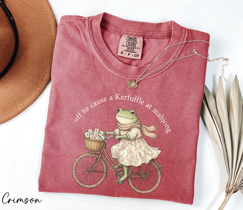 a red t-shirt with a cartoon image of a frog riding a bicycle, accompanied by the text "off to cause a Kerffuffle at mahjong".