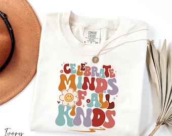 May include: Ivory-colored t-shirt with the words "CELEBRATE MINDS OF ALL KINDS" in a retro font. The design includes a smiling flower and other colorful graphics. A gold necklace and a brown hat are also visible.