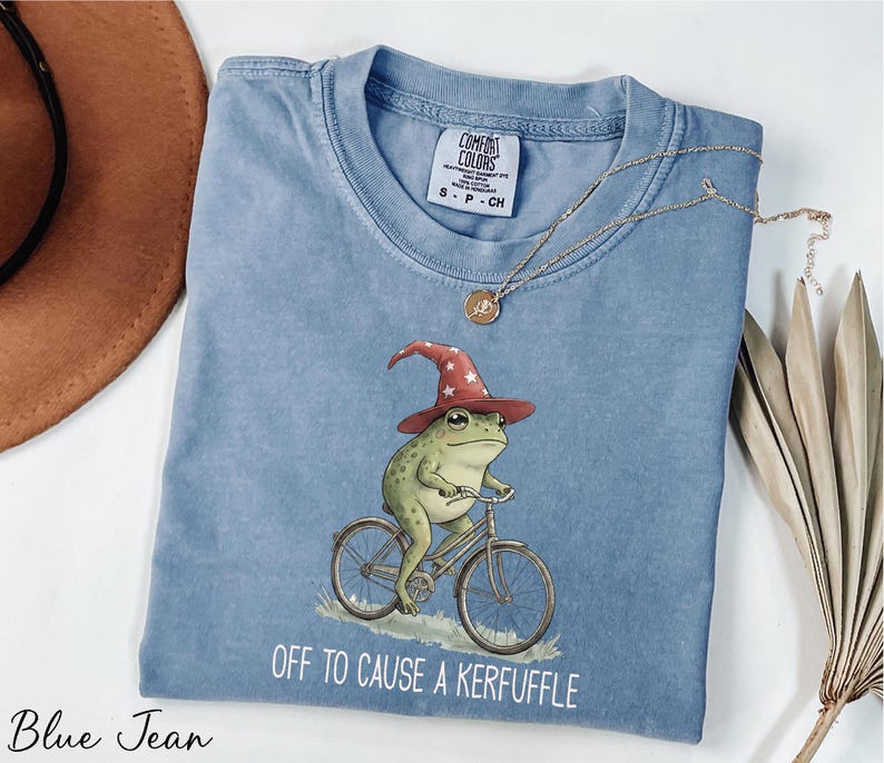 A blue t-shirt with a frog wearing a wizard's hat and riding a bicycle, accompanied by the text "Off to cause a Keruffle".