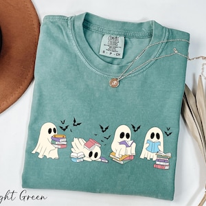 May include: Light green t-shirt with a graphic of four cartoon ghosts reading books. The ghosts are in different poses, with bats flying above them. The shirt has a crew neck and short sleeves. The text "Light Green" is at the bottom.