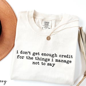 a white t-shirt with black text that reads "I don't get enough credit for the things I manage not to say", accompanied by a brown hat and a plant.