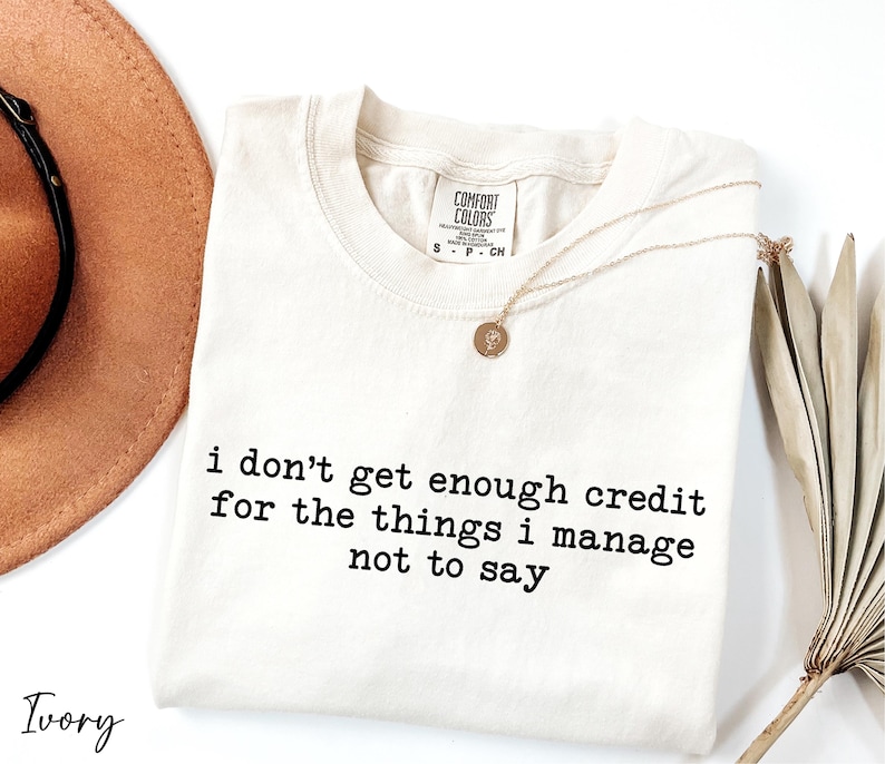a white t-shirt with black text that reads "I don't get enough credit for the things I manage not to say", accompanied by a brown hat and a plant.