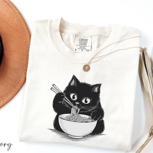 A white t-shirt with a black cat illustration and the word "Ivy" printed on it, along with a necklace and a hat.