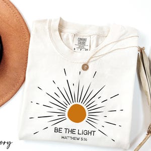 K&ouml;nnte beinhalten: Ein T-Shirt in Elfenbein mit einem schwarzen Sonnenstrahlen-Design und dem Text "BE THE LIGHT" mit "MATTHEW 5:14" darunter.