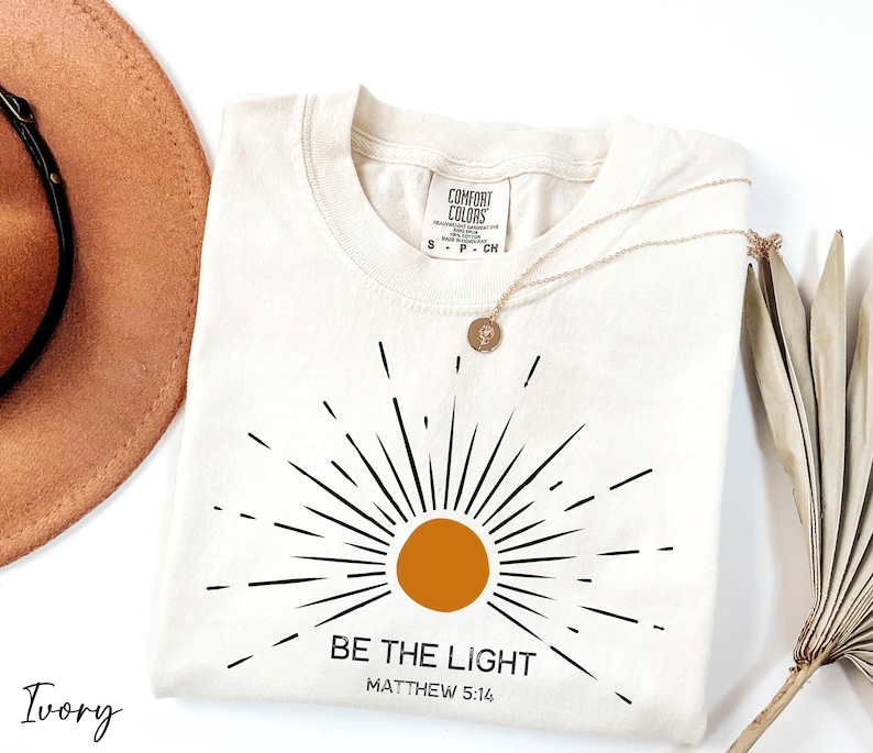 K&ouml;nnte beinhalten: Ein T-Shirt in Elfenbein mit einem schwarzen Sonnenstrahlen-Design und dem Text "BE THE LIGHT" mit "MATTHEW 5:14" darunter.