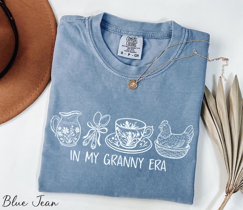 A blue t-shirt with a white graphic design, including a cup, spoon, and chicken, along with the text "In My Granny Era" printed on it. The t-shirt is laid out on a white surface, with a brown hat and dried leaves nearby.