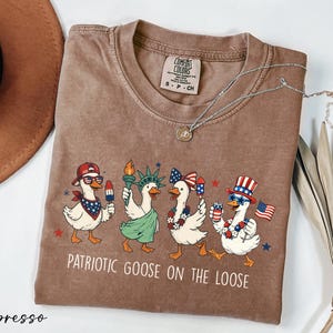 a brown t-shirt with a graphic design of four cartoon ducks dressed in patriotic outfits, including hats and scarves, dancing together. The text on the t-shirt reads "Patriotic Goose on the Loose".