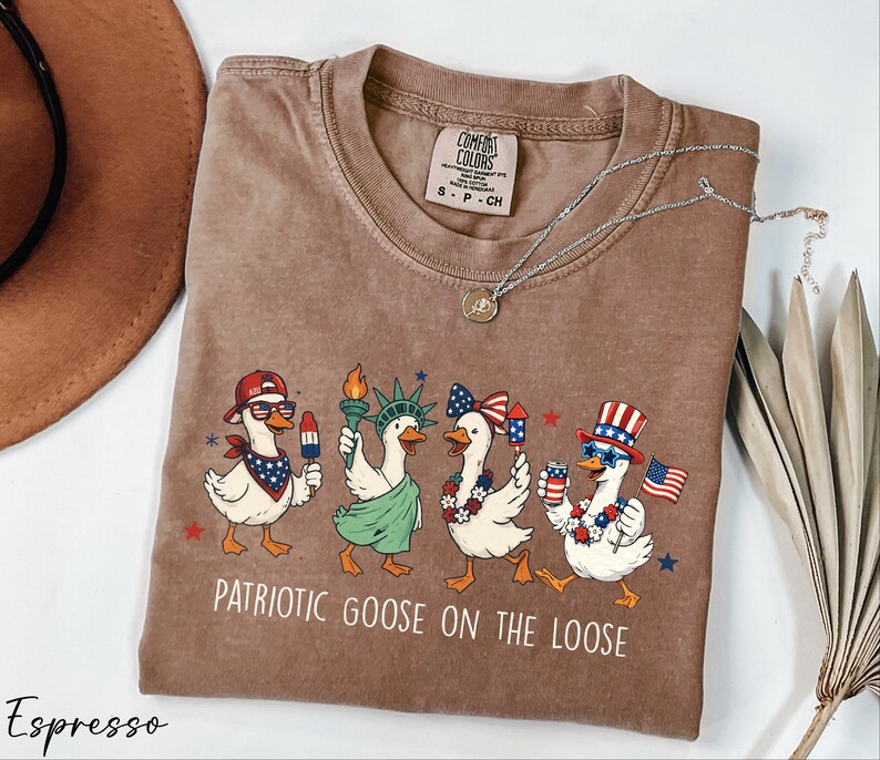 a brown t-shirt with a graphic design of four cartoon ducks dressed in patriotic outfits, including hats and scarves, dancing together. The text on the t-shirt reads "Patriotic Goose on the Loose".