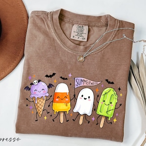 May include: An espresso-colored t-shirt featuring a graphic of four smiling characters: a purple ice cream cone with bat wings and a skull, a candy corn popsicle, a white ghost, and a green popsicle with stitches, holding a Summerween flag.