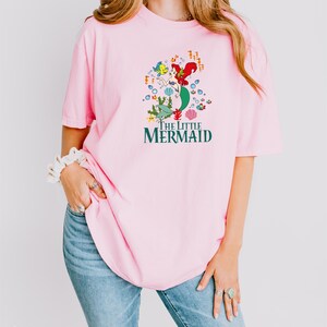 a woman wearing a pink shirt with the words the little mermaid on it