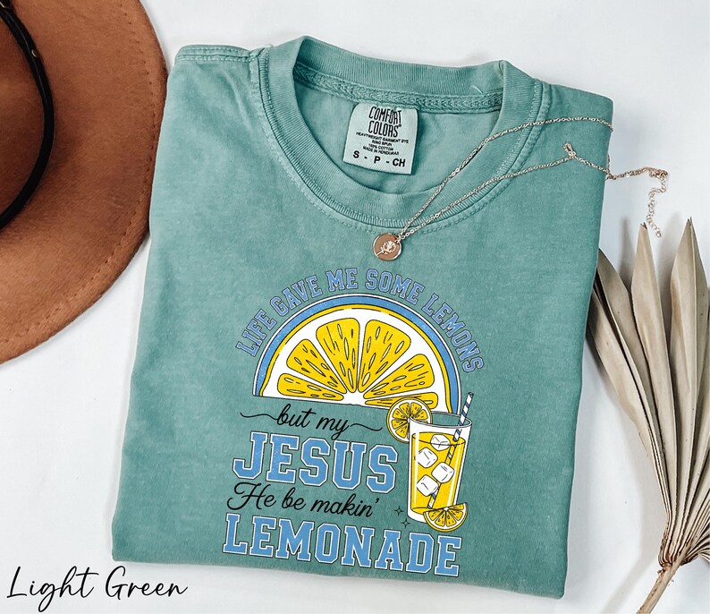 A light green t-shirt with a graphic design and text, along with a brown hat and some dried leaves, on a white background.