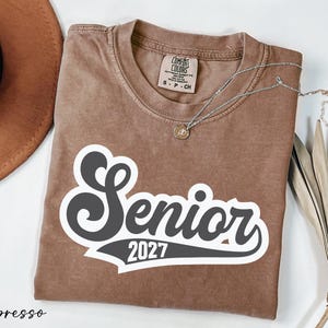 a brown t-shirt with the text "Senior 2027" printed on it, along with a necklace and a hat.