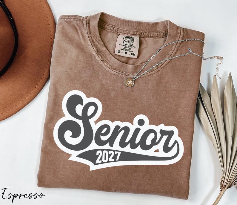 a brown t-shirt with the text "Senior 2027" printed on it, along with a necklace and a hat.