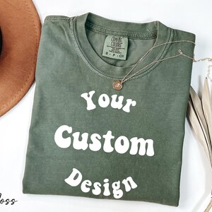 Op de afbeelding: Een mosgroen T-shirt met de tekst "Your Custom Design" in wit, retro-stijl lettertype. Het shirt is gevouwen en ligt plat op een wit oppervlak.