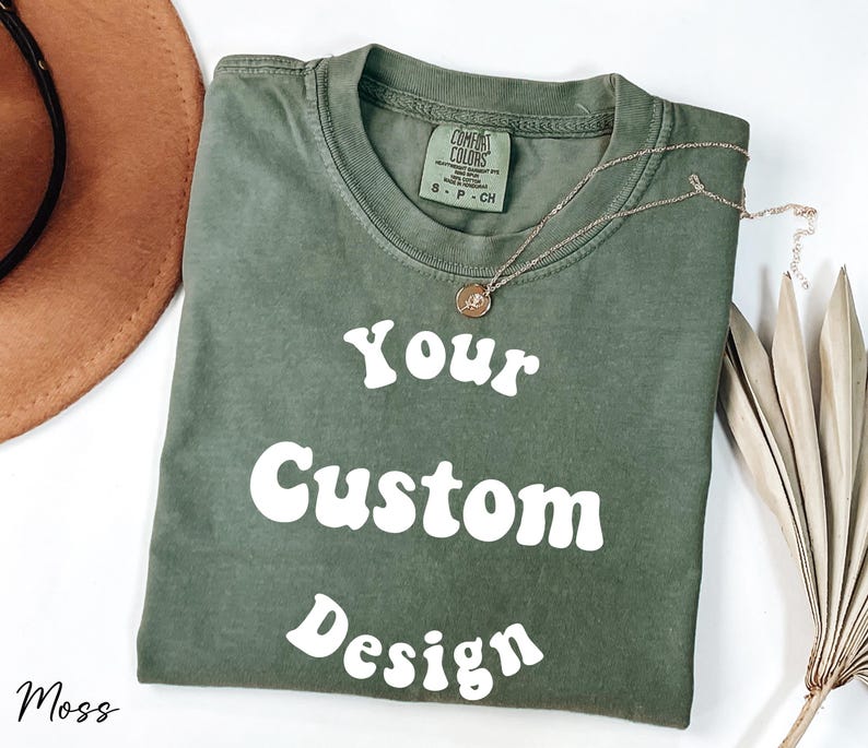 Op de afbeelding: Een mosgroen T-shirt met de tekst "Your Custom Design" in wit, retro-stijl lettertype. Het shirt is gevouwen en ligt plat op een wit oppervlak.