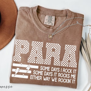 a brown t-shirt with a checkered pattern and the word "PARA" printed on it, along with some text below the word. The text reads "Some days I rock it, some days it rocks me either way we rockin' it" and is located in the center of the t-shirt. The t-shirt is laid out on a white surface, with a hat and some leaves visible in the background.
