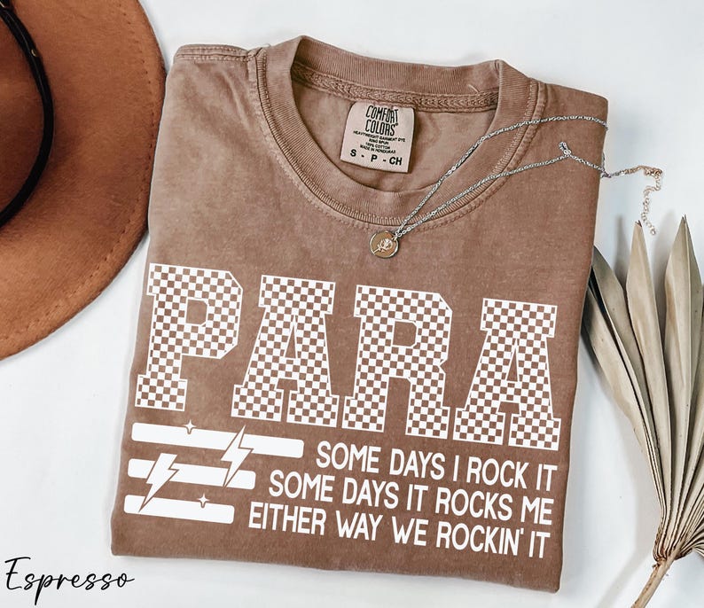 a brown t-shirt with a checkered pattern and the word "PARA" printed on it, along with some text below the word. The text reads "Some days I rock it, some days it rocks me either way we rockin' it" and is located in the center of the t-shirt. The t-shirt is laid out on a white surface, with a hat and some leaves visible in the background.