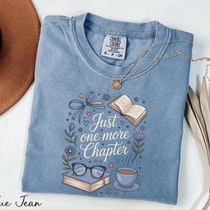 A blue t-shirt with a printed design, a pair of glasses, a book, a cup, and a plant, along with a necklace and a hat, all laid out on a white surface.