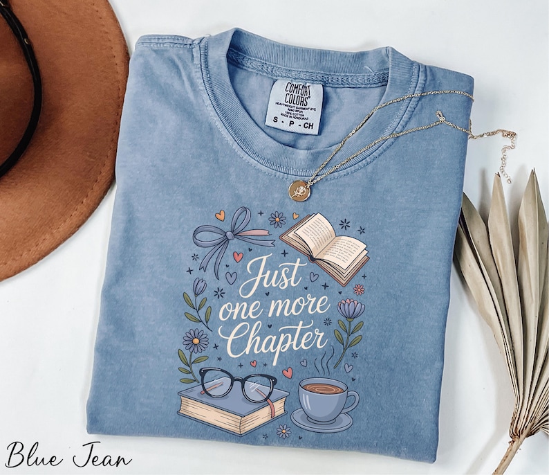 A blue t-shirt with a printed design, a pair of glasses, a book, a cup, and a plant, along with a necklace and a hat, all laid out on a white surface.