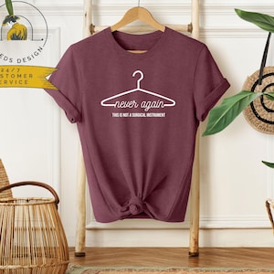 May include: A maroon t-shirt with the words "never again" and "this is not a surgical instrument" printed in white. The design features a hanger graphic. The shirt is hanging on a wooden hanger.