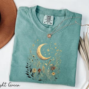 A light green t-shirt with a moon and starry night sky design, along with a necklace and a brown hat, are displayed on a white surface.