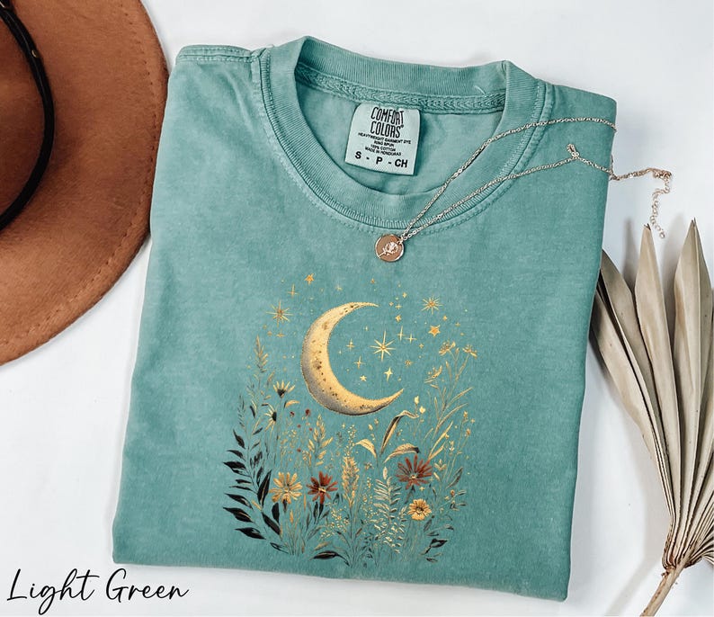 A light green t-shirt with a moon and starry night sky design, along with a necklace and a brown hat, are displayed on a white surface.