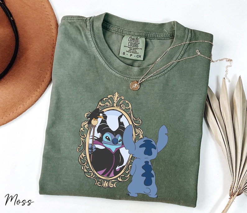 A green t-shirt with a design featuring a character from the Disney movie "Stitch" and a mirror, along with a necklace and a hat.