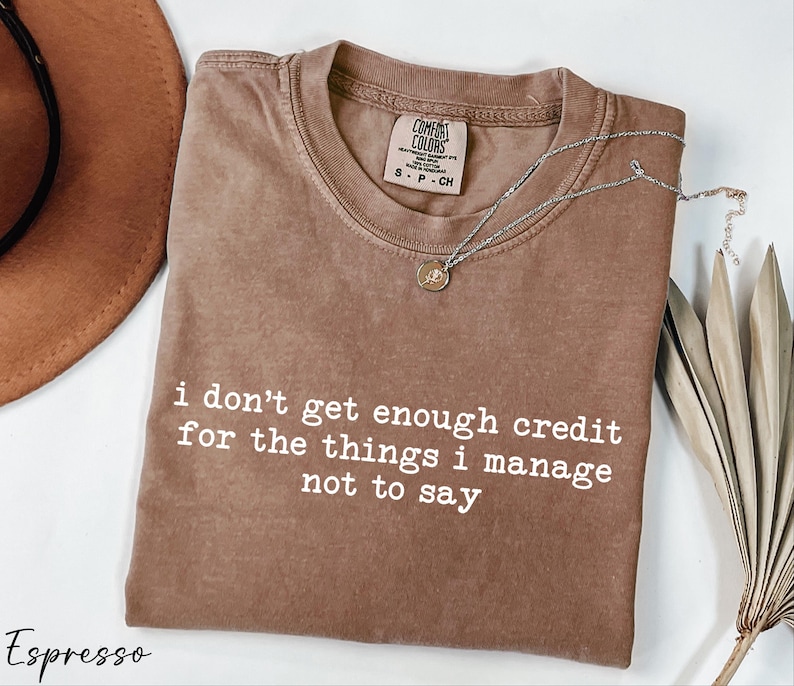 a brown t-shirt with a humorous text printed on it, along with a hat, a necklace, and some dried leaves, all arranged on a white background.