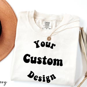 Op de afbeelding: Een ivoorwit T-shirt met de tekst "Your Custom Design" in een retro-lettertype.