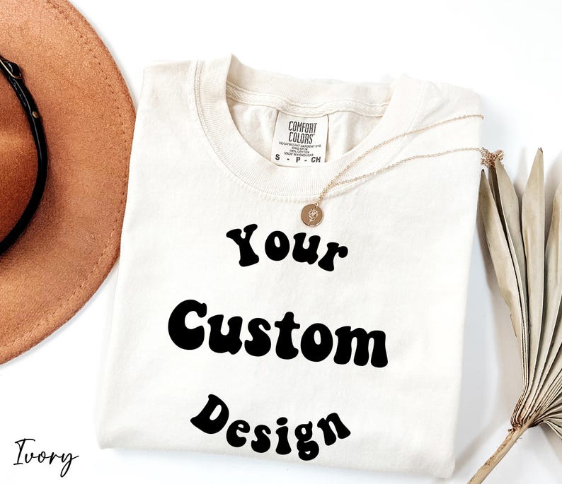 Op de afbeelding: Een ivoorwit T-shirt met de tekst "Your Custom Design" in een retro-lettertype.