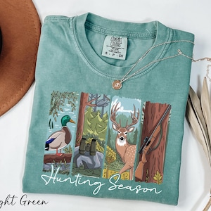 A light green t-shirt with a graphic design depicting various hunting scenes, including a duck, deer, and rifle, along with the text "Hunting Season".