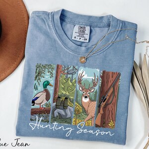 A blue t-shirt with a graphic design depicting a duck, deer, and bird in a forest setting, along with the text "Hunting Season". The t-shirt is laid out on a white surface, with a brown hat and some dried leaves nearby.
