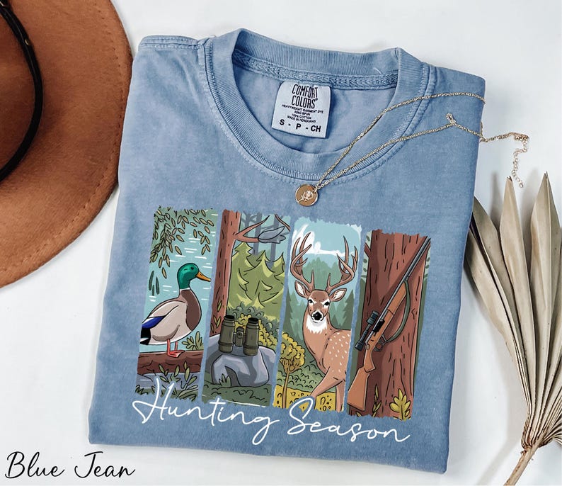 A blue t-shirt with a graphic design depicting a duck, deer, and bird in a forest setting, along with the text "Hunting Season". The t-shirt is laid out on a white surface, with a brown hat and some dried leaves nearby.