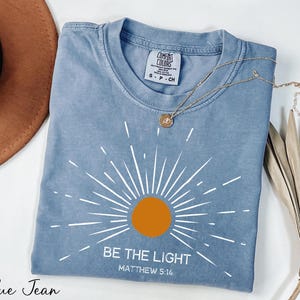 K&ouml;nnte beinhalten: Ein blaues Jeans-T-Shirt mit einem wei&szlig;en Sonnenstrahlen-Design und dem Text "Sei das Licht" mit dem Bibelvers "Matth&auml;us 5:16".