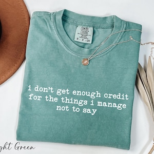 A light green t-shirt with a quote printed on it, along with a brown hat and some dried leaves, all placed on a white surface.