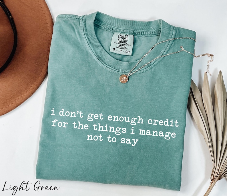 A light green t-shirt with a quote printed on it, along with a brown hat and some dried leaves, all placed on a white surface.