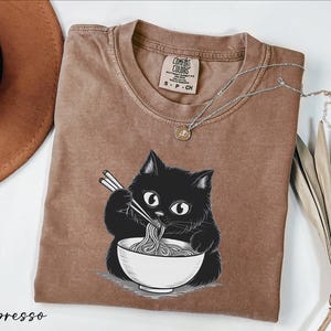 a brown t-shirt with a black cat graphic, holding a bowl of noodles and eating with chopsticks.