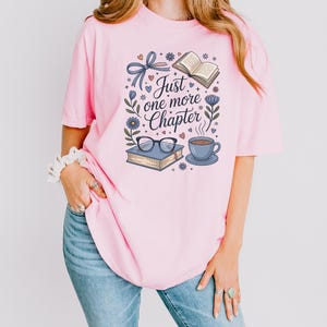 A woman wearing a pink t-shirt with a graphic design featuring a book, glasses, and a cup of coffee, along with the text "Just one more Chapter".