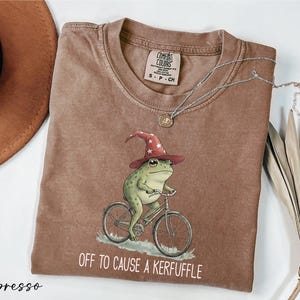 a brown t-shirt with a whimsical illustration of a frog riding a bicycle, wearing a wizard's hat. The text on the t-shirt says "Off to cause a Kerffle".